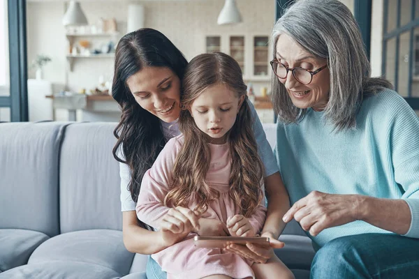 Klein meisje met moeder en grootmoeder met behulp van smartphone tijdens het zitten op de bank thuis — Stockfoto