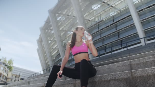 Belle jeune femme en vêtements de sport boire de l'eau tout en étant assis à l'extérieur — Video