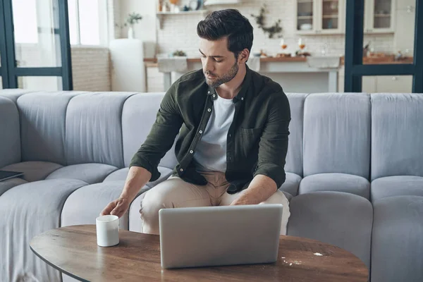 Snygg ung man som använder laptop och dricker kaffe när han sitter på soffan hemma — Stockfoto