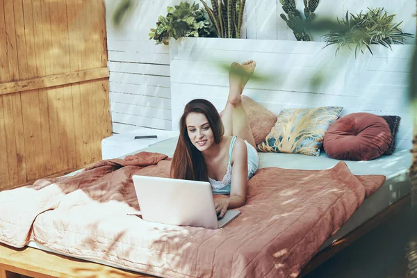 Όμορφη νεαρή χαμογελαστή γυναίκα χρησιμοποιώντας φορητό υπολογιστή, ενώ βρίσκεται στο κρεβάτι — Φωτογραφία Αρχείου