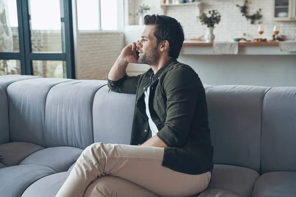 Snygg ung man pratar i smart telefon och tittar bort medan du sitter på soffan — Stockfoto