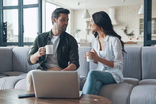 Bela jovem casal bebendo café e olhando no laptop enquanto sentado no sofá em casa — Fotografia de Stock