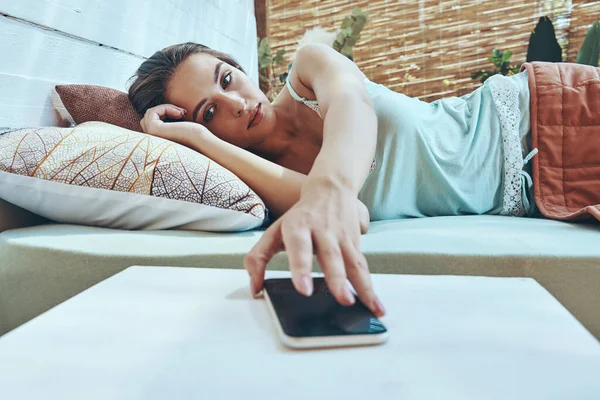 Gyönyörű fiatal nő veszi a mobiltelefonját, miközben fekszik a házi ágyban — Stock Fotó