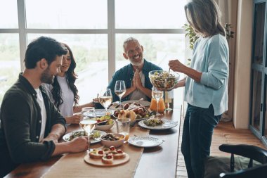 Modern bir dairede oturmuş yemek yiyip gülümseyen çok nesilden güzel bir aile.