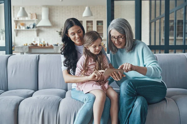 Klein meisje met moeder en grootmoeder met behulp van smartphone tijdens het zitten op de bank thuis — Stockfoto
