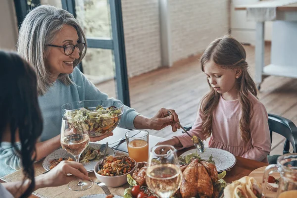 Hermosa mujer mayor poniendo ensalada para niña mientras cenan juntos — Foto de Stock