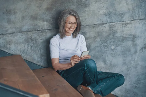 Senior hermosa mujer usando el teléfono inteligente y sonriendo mientras está sentado en las escaleras en el interior — Foto de Stock