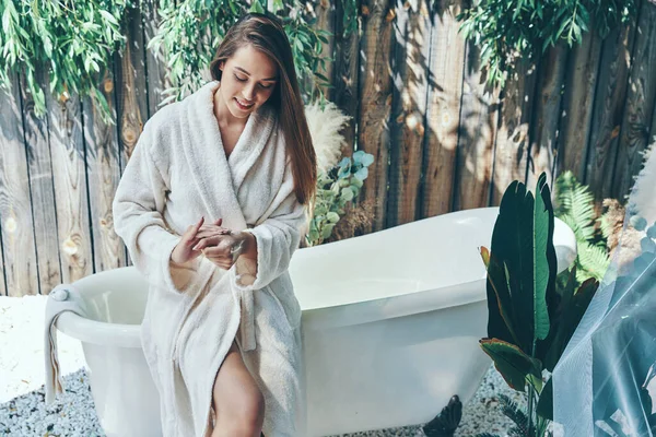 Hermosa joven sonriente crema hidratante para las manos mientras se inclina en la bañera al aire libre —  Fotos de Stock