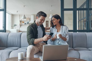 Yakışıklı genç kız arkadaşına akıllı telefonunu gösteriyor ve evde kanepede otururken gülümsüyor.