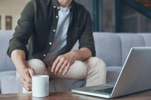 Primer plano del hombre joven usando el ordenador portátil y beber café mientras está sentado en el sofá en casa — Foto de Stock