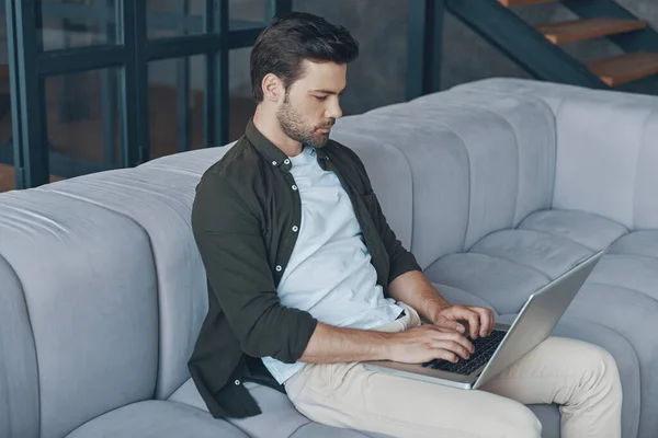 Hombre joven guapo usando el ordenador portátil mientras está sentado en el sofá en casa — Foto de Stock