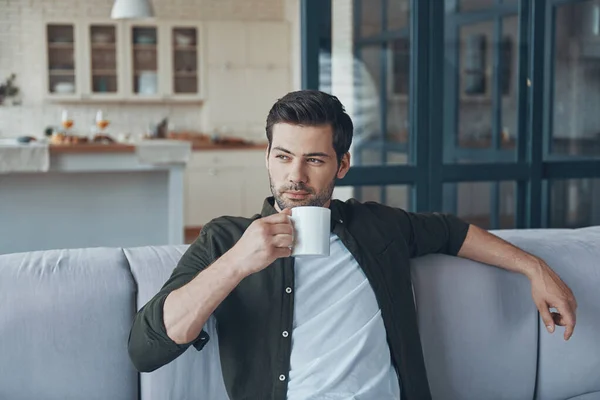 Handsome young man holding cup with drink while sitting on the sofa at home — Fotografia de Stock