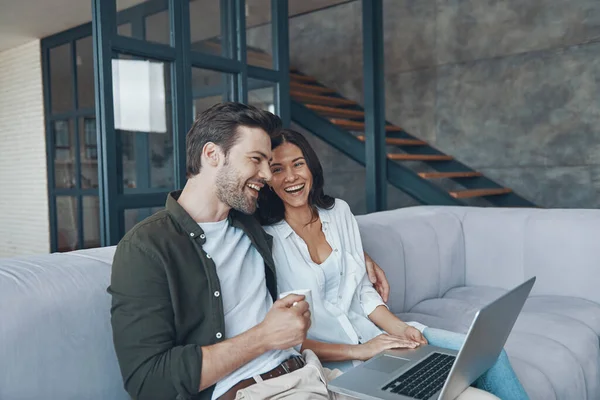 Mooi jong stel drinken van koffie en kijken op de laptop terwijl zitten op de bank thuis — Stockfoto