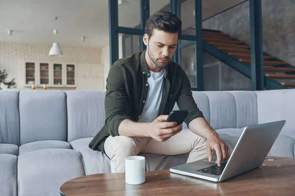 Snygg ung man som använder laptop när du sitter på soffan hemma — Stockfoto