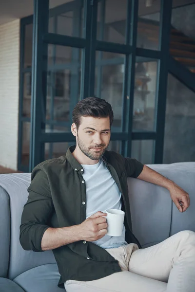 Красивий молодий чоловік тримає чашку з напоєм, сидячи на дивані вдома — стокове фото