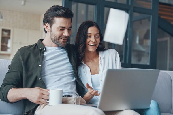 Красивая молодая пара пьет кофе и смотрит на ноутбук, сидя дома на диване