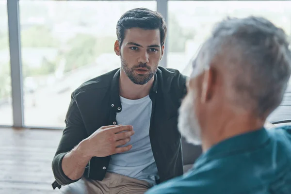 Handsome young man talking to his senior father while spending time at home together — Stock Photo, Image