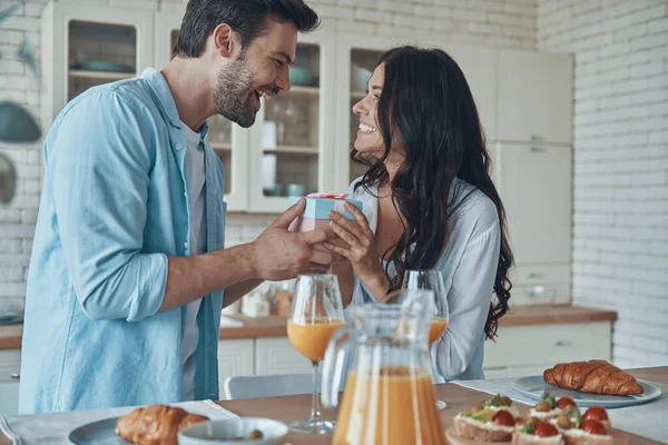 Ung man ger en presentförpackning till sin flickvän samtidigt som han äter frukost i hushållet kök — Stockfoto