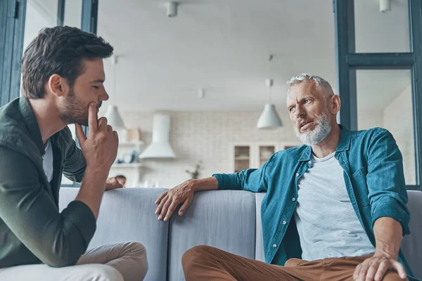 Handsome young man talking to his senior father while spending time at home together — Stock Photo, Image