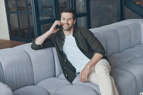 Handsome young man talking on smart phone and looking away while sitting on the sofa at home — Fotografia de Stock