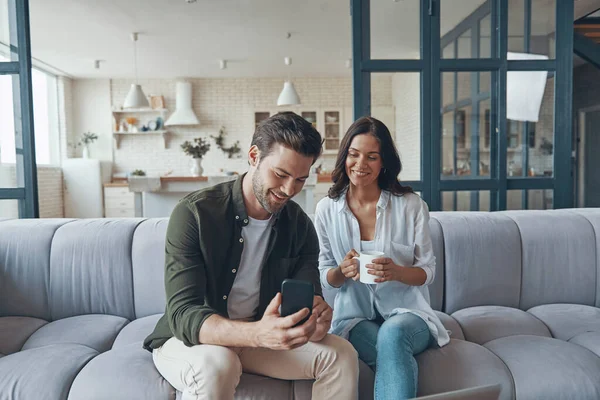 Beautiful young couple drinking coffee and communicating while sitting on the sofa at home — Fotografia de Stock
