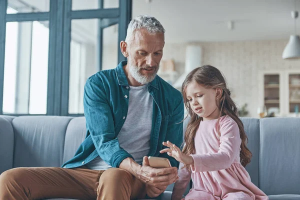 Linda niña usando el teléfono inteligente mientras pasa tiempo con el abuelo y en casa —  Fotos de Stock