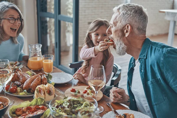 Happy little girl feeding her grandfather and smiling while having dinner with family — Stock Photo, Image