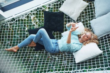 Evdeki büyük hamakta dinlenirken akıllı telefon kullanan güzel bir genç kadın manzarası.