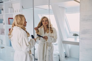 Evde aynanın önünde dikilirken saçlarını tarayan bornozlu çekici genç bir kadın.