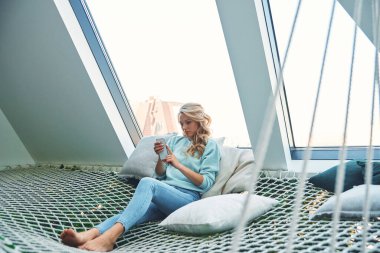 Güzel genç bayan evde büyük bir hamakta dinlenirken akıllı telefon kullanıyor.