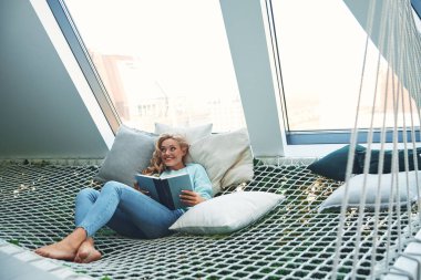 Güzel genç bir kadın kitap okuyor ve gülümsüyor evde büyük bir hamakta dinlenirken.