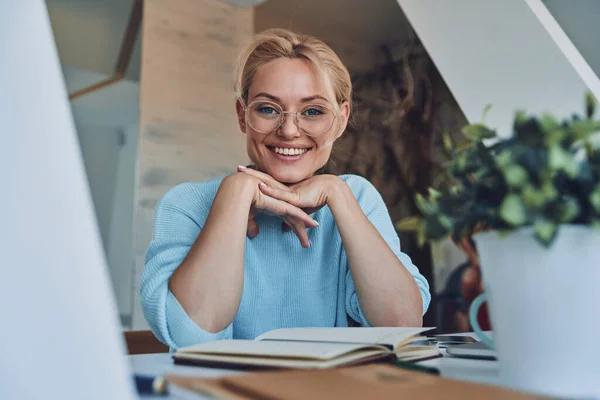 Mooie jonge vrouw kijken naar camera en glimlachen terwijl zitten op haar werkplek in kantoor — Stockfoto
