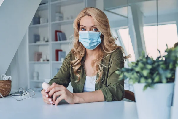 Attractive young woman in protective face mask sitting at the desk in office and looking at camera — Stock Photo, Image