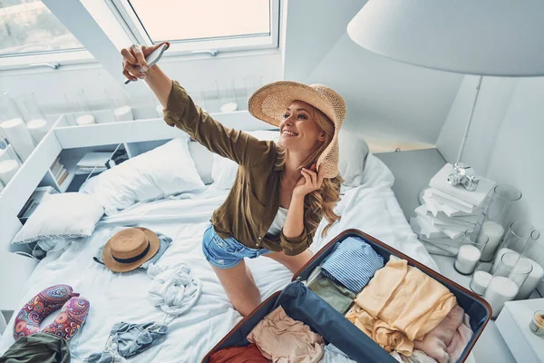 Vista superior de una hermosa joven sonriente haciendo selfie mientras empaca el equipaje en casa —  Fotos de Stock