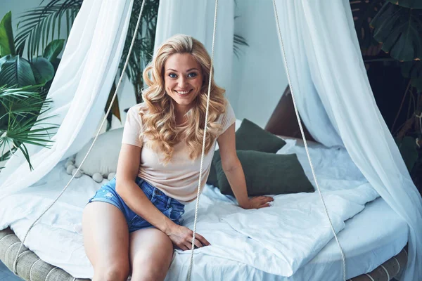 Gyönyörű fiatal nő néz kamera és mosolyog, miközben ül az ágyon otthon — Stock Fotó
