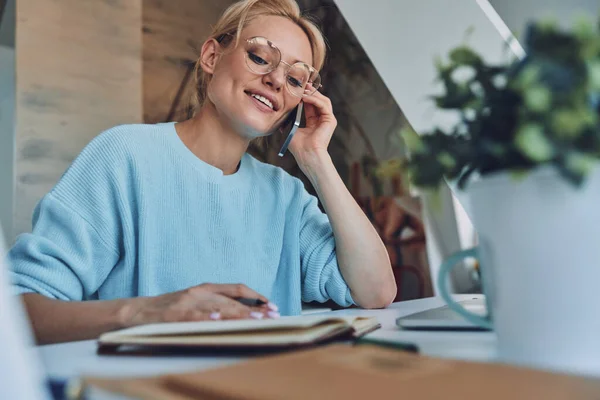 Vacker ung kvinna skriver något i anteckningsblock och talar på mobiltelefon när du arbetar på kontoret — Stockfoto