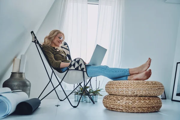 Aantrekkelijke jonge vrouw met behulp van laptop terwijl zitten in comfortabele stoel thuis — Stockfoto