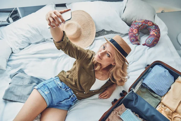 Vista superior de una hermosa joven sonriente haciendo selfie mientras empaca el equipaje en casa —  Fotos de Stock