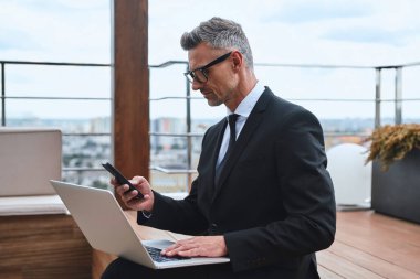 Diz üstü bilgisayarda çalışan ve çatıdaki terasta dururken cep telefonu kullanan kendinden emin olgun bir adam.