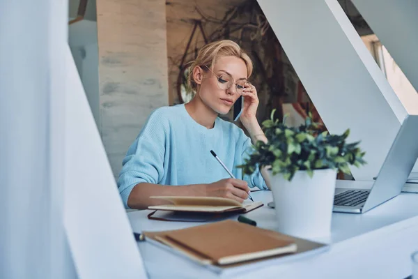 Gyönyörű fiatal nő ír valamit jegyzetfüzetbe, és beszél mobiltelefonnal, miközben dolgozik az irodában — Stock Fotó