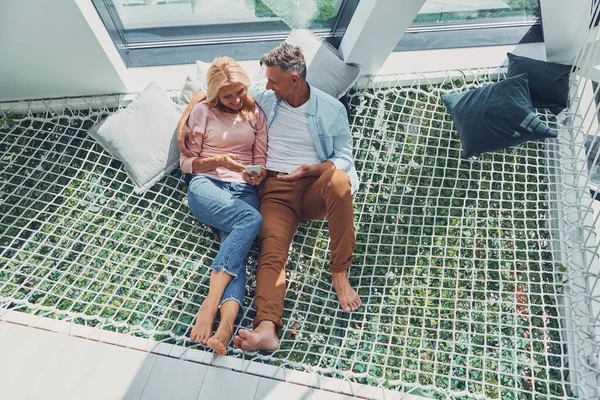 Gyönyörű érett pár vizsgálja okos telefon és mosolyog, miközben pihentető nagy függőágy otthon együtt — Stock Fotó