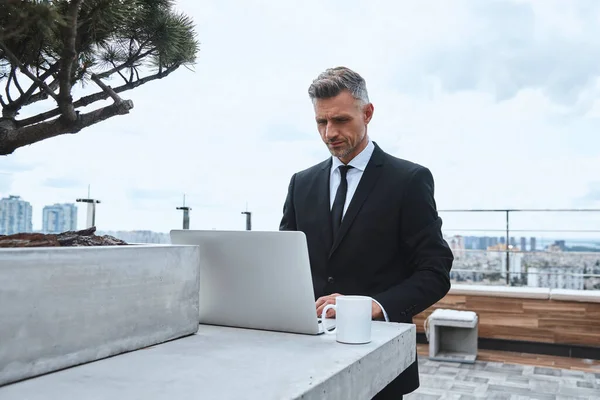 Sebevědomý dospělý muž pracující na notebooku, zatímco stojí na střešní terase — Stock fotografie