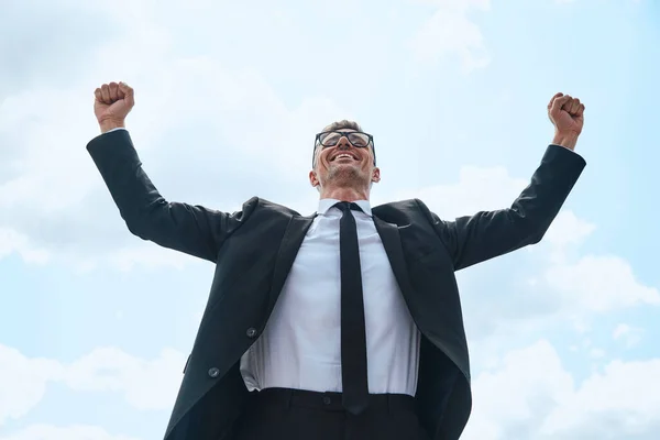 Heureux homme mature en costume complet gardant les bras levés tout en se tenant devant le ciel bleu à l'extérieur — Photo