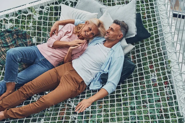 Vista superior de la hermosa pareja madura sonriendo y comunicándose mientras se relaja en la hamaca grande en casa juntos —  Fotos de Stock