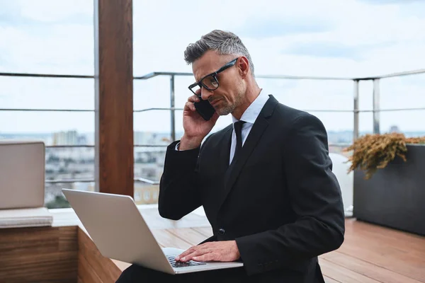 Homme mature confiant travaillant sur un ordinateur portable et utilisant un téléphone portable tout en se tenant debout sur le toit-terrasse — Photo