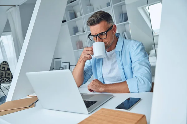Homme mature confiant travaillant sur un ordinateur portable et buvant du café tout en étant assis au bureau — Photo