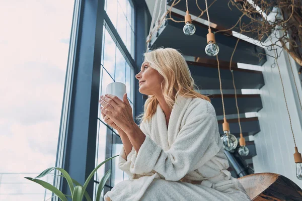 Gyönyörű érett nő fürdőköpenyben kávét iszik és mosolyog, miközben otthon ül az ablak mellett. — Stock Fotó