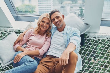 Mutlu, olgun bir çift, evde büyük bir hamakta dinlenirken gülümseyip kameraya bakıyor.