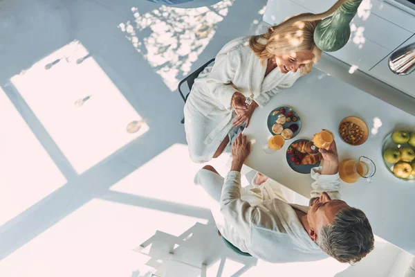 Vista superior do belo casal maduro em roupões de banho desfrutando de café da manhã juntos enquanto passam tempo na cozinha doméstica — Fotografia de Stock