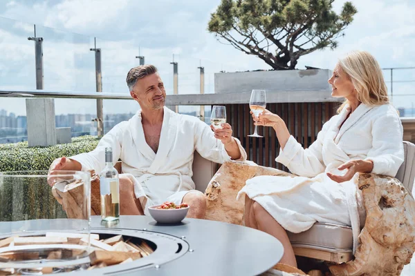Vackra mogna par i badrockar njuter av frukt och vin samtidigt som de kopplar av i lyxhotell utomhus — Stockfoto